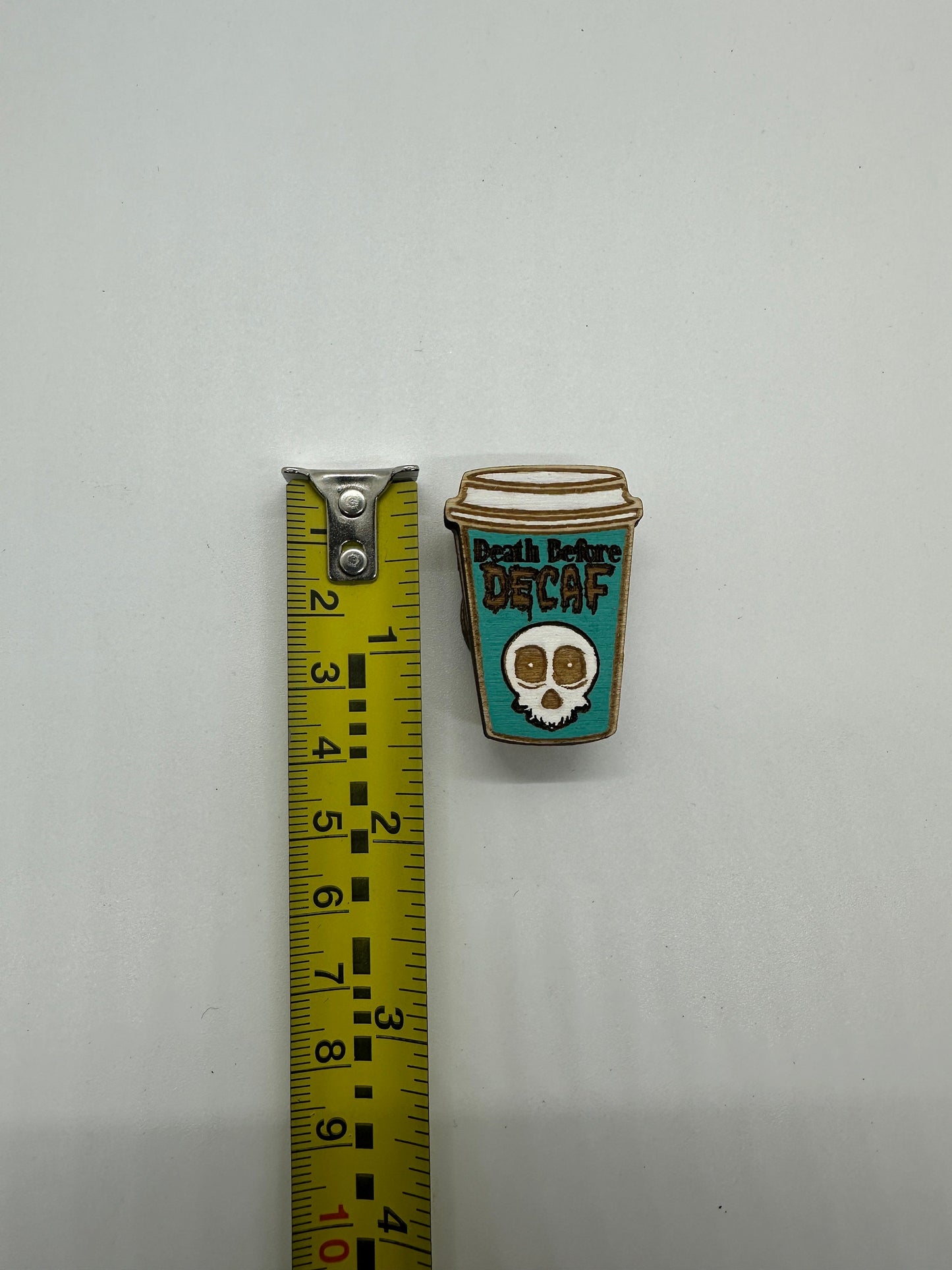 Pin with 'Death Before Decaf' design on a white background with a ruler for scale.