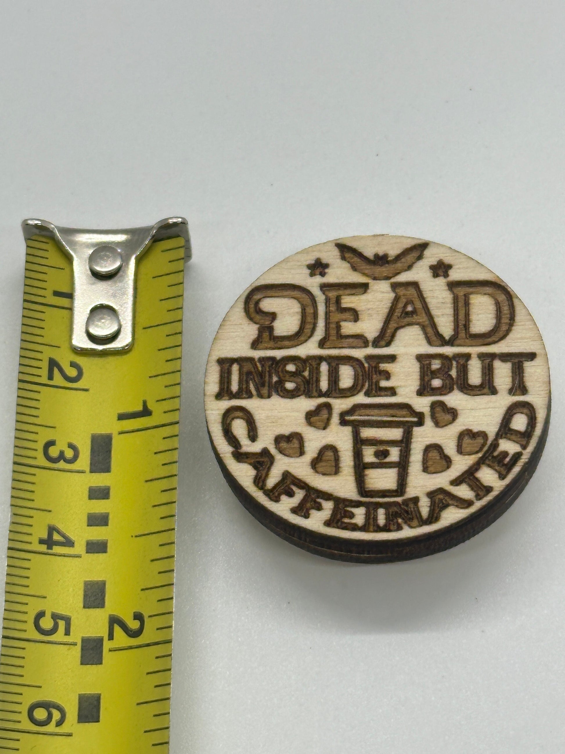 Wooden round sign with 'Dead Inside But Caffeinated' text and a coffee cup graphic, next to a tape measure for scale.