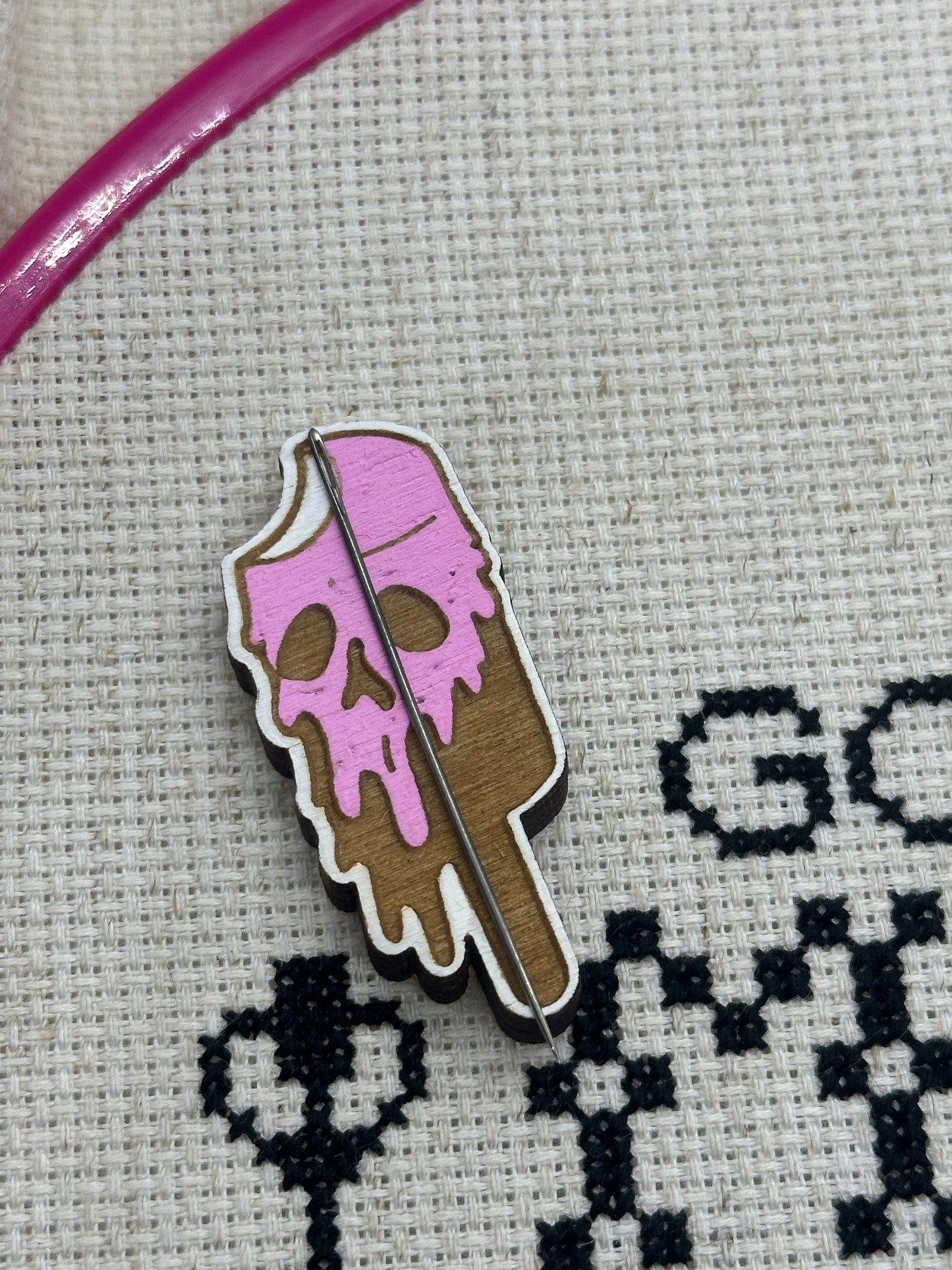Pink skull-shaped ice cream cone pin on a cross-stitched fabric background