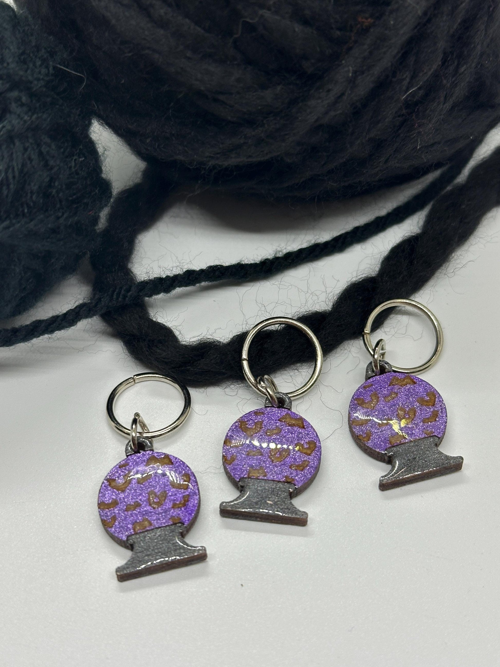 Three purple and brown patterned knitting stitch markers with a ball of black yarn in the background.